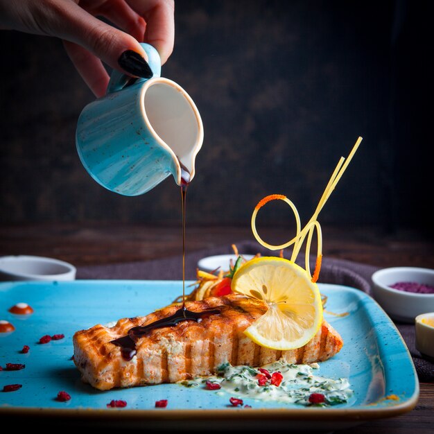 Close-up grilled fish, hand pouring sauce with garnish, lemon, sauce and spices on a blue plate on a dark wooden table