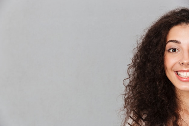 Close up half face portrait of attractive curly woman with ring in nose posing smiling with perfect white teeth over grey wall