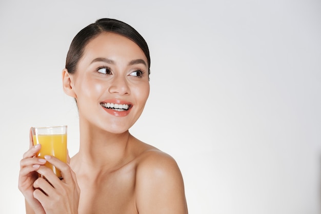 Close up of half-naked gentle woman with healthy fresh skin looking away and holding orange juice from transparent glass, isolated over white wall