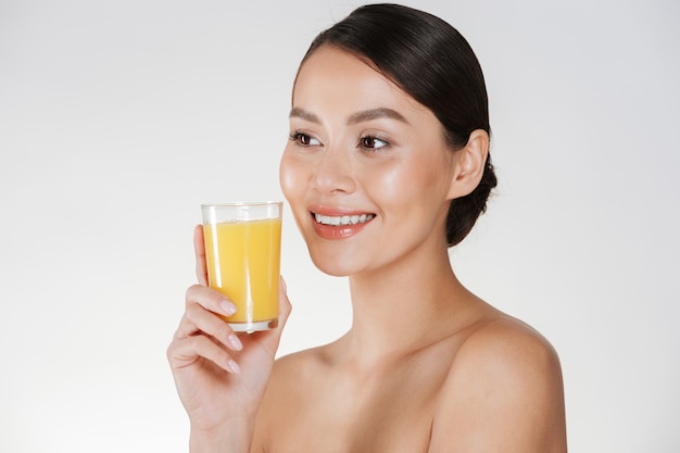 Close up of half-naked lady with healthy fresh skin and broad smile drinking orange juice from transparent glass, isolated over white wall