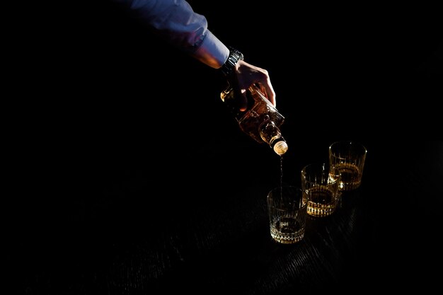Крупным планом рука, держащая бутылку, наливает напиток в стакан в черном темном пространстве Концепция празднования и мальчишника коктейлей бармена