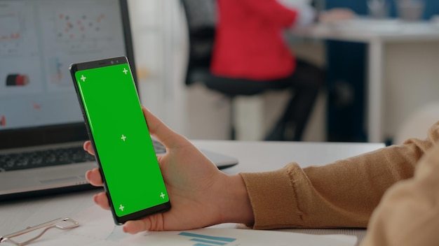 携帯電話で垂直に緑色の画面を持っている手のクローズアップ。空白の背景とビジネスオフィスで分離されたクロマキーテンプレートのモックアップで作業している人。モックアップ表示