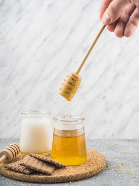 Close-up of hand holding honey dipper over honey pot with biscuits on cork coaster