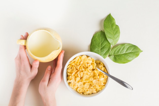 Close-up of hand holding milk pitcher; cornflakes bowl and leaves on white background