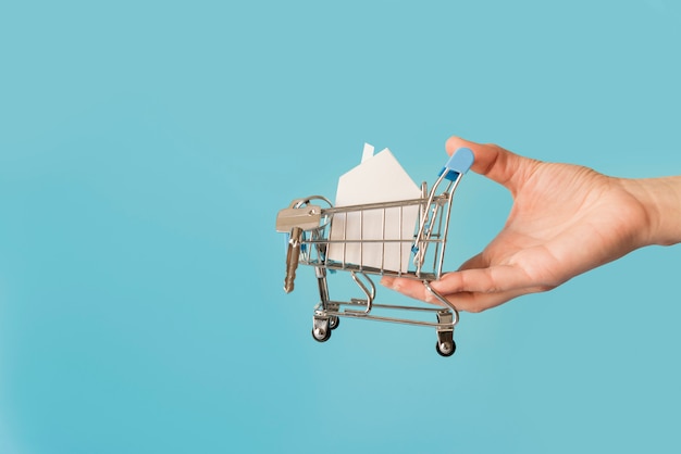 Close-up of hand holding miniature shopping cart with paper house and keys against blue background
