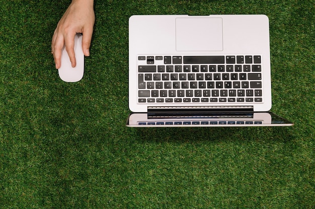 Primo piano della mano che tiene il mouse con il computer portatile aperto sul contesto di erba finta