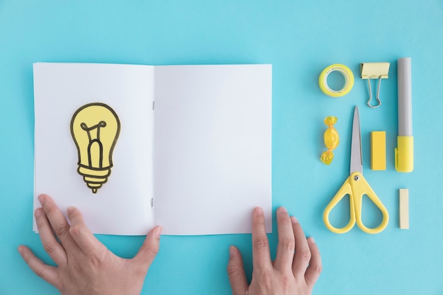 Close-up of hand holding page with light bulb and stationery on blue background