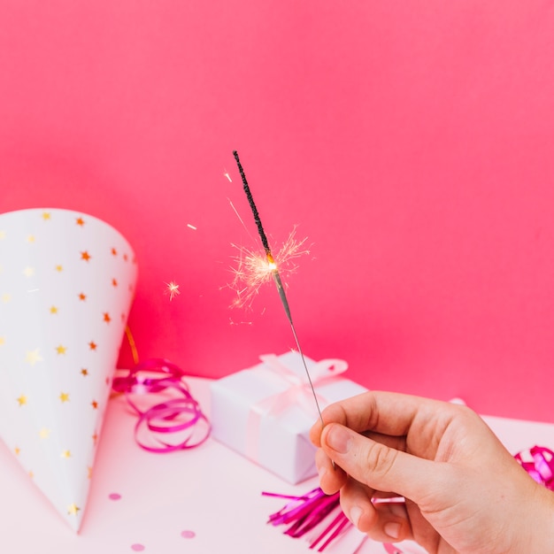 Close-up of hand holding sparkler on birthday celebration against pink background