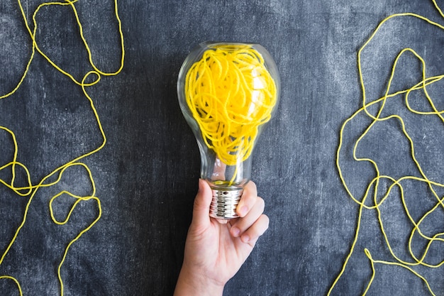 Close-up of hand holding transparent light bulb with yellow yarn on blackboard