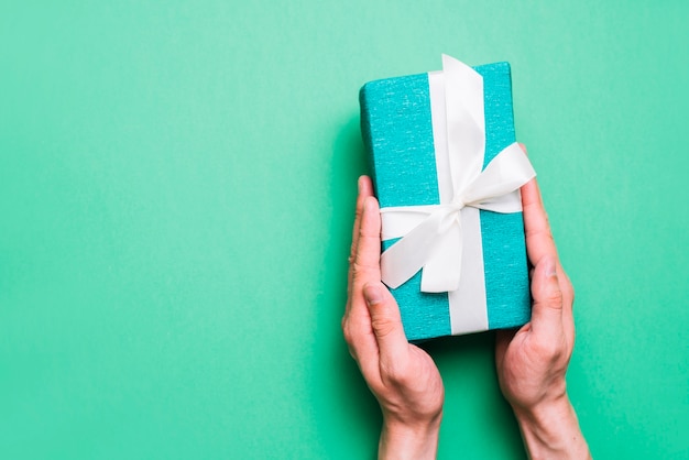 Close-up of hand holding wrapped gift box with white ribbon against green background