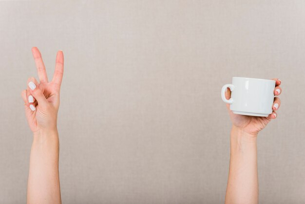 Close-up of hand making peace gesture and white cup against colored backdrop