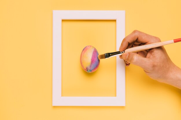 Close-up of hand painting the easter egg with brush inside the frame on yellow background