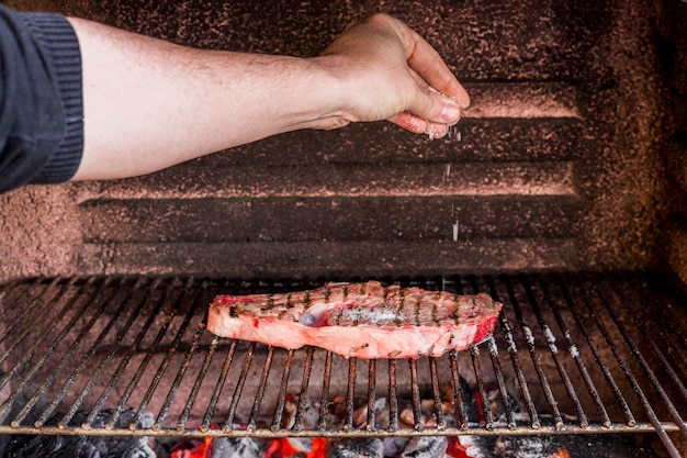 Close-up of hand sprinkling salt on grilled beef in barbecue