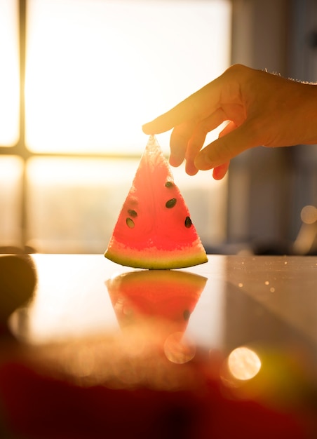 Close-up of hand touching the slice of watermelon on desk against sunlight