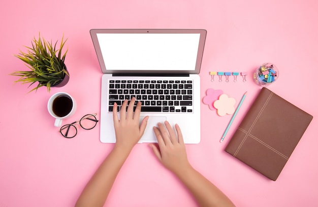 Close-up of hand typing on laptop with stationeries and coffee cup on pink backdrop