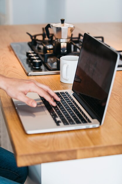 Close-up of hand typing on laptop over the wooden kitchen counter with coffee cup