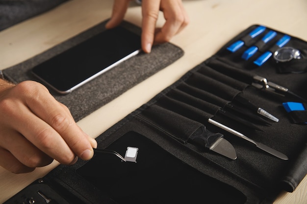 Close up on hand with pincher tool holding sim-card slot with nano sim above black magnet plate on portable tookit for electronic repeirment service