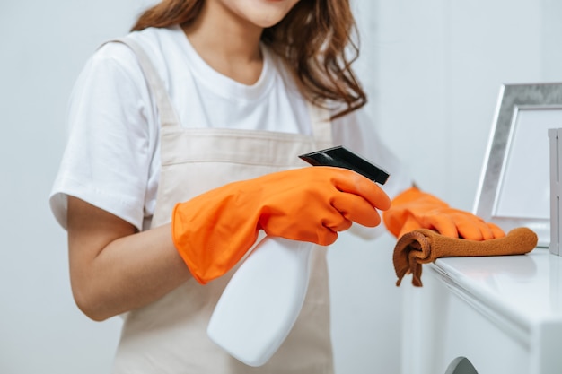 La mano ravvicinata della giovane donna governante in guanti di gomma usa la soluzione detergente in un flacone spray su mobili bianchi e usa un panno per pulirlo