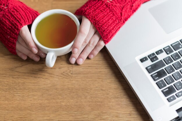 Close-up of hands holding a cup of tea next to a laptop