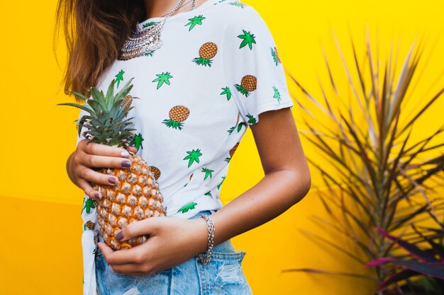 Close-up hands holding pineapple tanned skin of attractive woman on vacation wearing straw hat barefoot in denim shorts printed t-shirt summer fashion