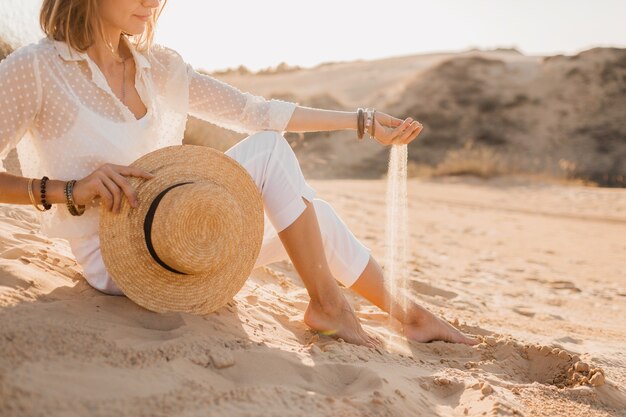 Close-up hands with sand of stylish beautiful woman in desert in white outfit holding straw hat on sunset