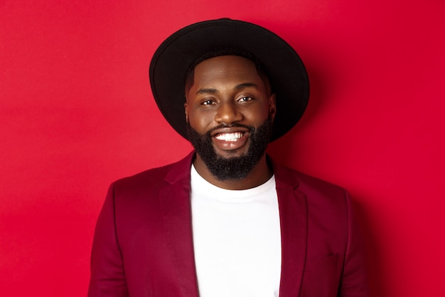 Close-up of handsome african american man with beard, wearing party blazer and stylish hat, smiling at camera, red background