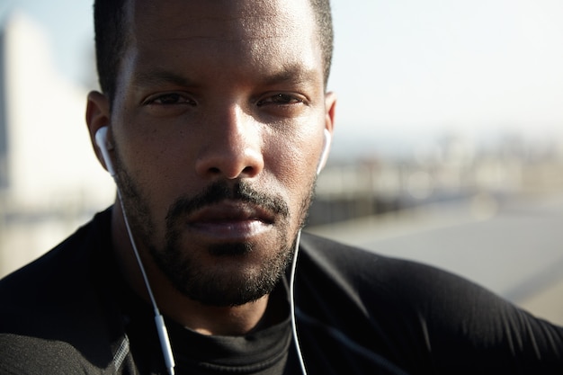 Close up of handsome African athlete with healthy tanned skin and confident look, wearing black sportswear, squinting his eyes while resting outdoors, listening to his favourite music with earphones