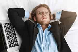 Free photo close up of handsome bearded businessman in fashionable suit lying on back with hands under head with laptop near him, looking upside, thinking about tomorrow's meeting.