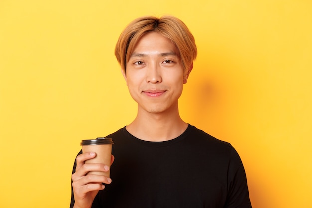 Close-up of handsome blond asian man drinking coffee and smiling pleased, standing over yellow wall