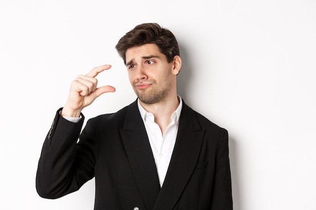 Close-up of handsome businessman in trendy suit, showing something small with disappointment, standing against white background.