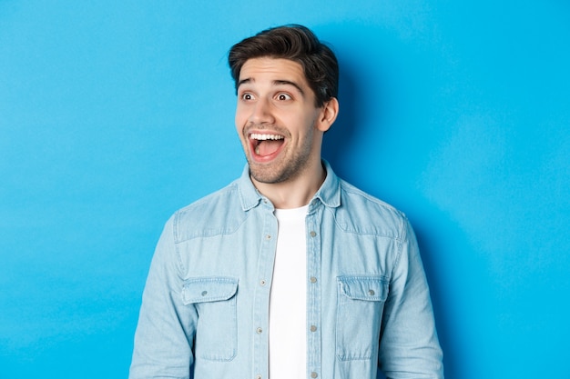 Close-up of handsome caucasian male model looking amazed left, checking out advertisement, standing over blue background.