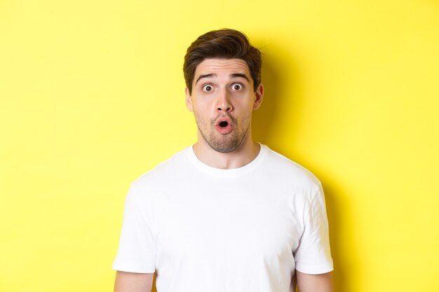 Close-up of handsome caucasian man saying wow, looking impressed, standing against yellow background.