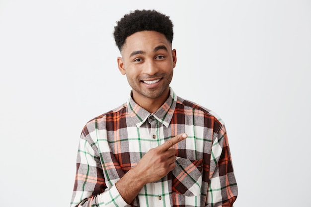 Close up of handsome cheerful tan-skinned man with afro hairstyle in casual checkered shirt smiling brightly, pointing aside with index finger o white wall. Copy space