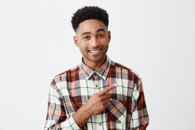 Close up of handsome cheerful tan-skinned man with afro hairstyle in casual checkered shirt smiling brightly, pointing aside with index finger o white wall. Copy space