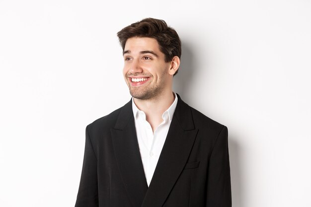 Крупным планом красивый мужчина-предприниматель в костюме, глядя влево и улыбаясь, стоя на белом фоне.