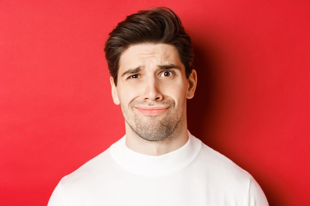 Close-up of handsome man feeling uncomfortable, grimacing and looking at something displeasing, standing over red background