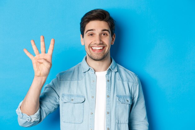 웃는 잘생긴 남자의 클로즈업, 4번 손가락을 보여주는 파란색 배경 위에 서 있는