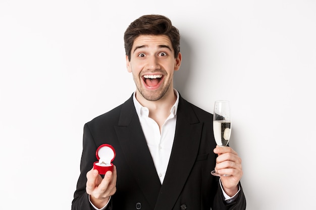 Close-up of handsome man in suit, making a proposal, giving engagement ring and raising glass of champagne, standing against white background