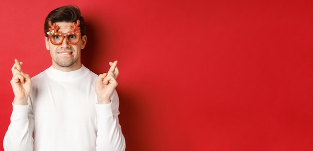 Close-up of handsome nervous guy in party glasses, making a wish, crossing fingers for good luck and looking with hope at camera, standing over red background.