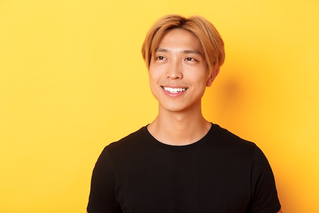 Close-up of handsome stylish korean guy with fair hair, looking dreamy and hopeful upper left corner, smiling pleased, standing yellow wall