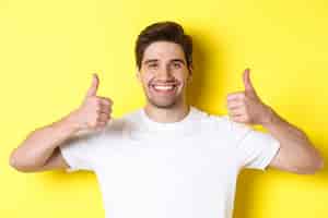 Free photo close-up of handsome young man showing thumbs up, approve and agree, smiling satisfied, standing over yellow background.