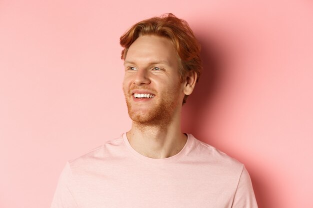Close up of handsome young man with beard and red hair, looking left and smiling delighted, standing confident over pink background