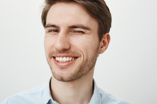 Close-up of handsome young man with white teeth smiling pleased, winking cheeky