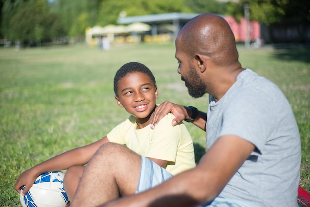 Крупным планом счастливый афро-американский отец разговаривает со своим сыном. Красивый мужчина сидит на земле, трогательно улыбаясь мальчику плечом, глядя друг на друга. Концепция воспитания, досуга и активного отдыха