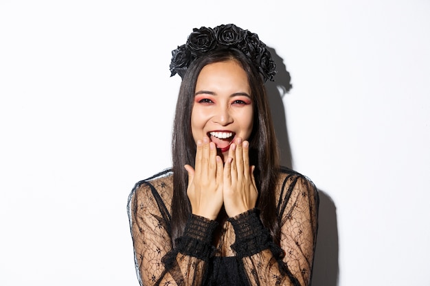 Close-up of happy asian woman celebrating halloween in witch costume and laughing, standing over white background.