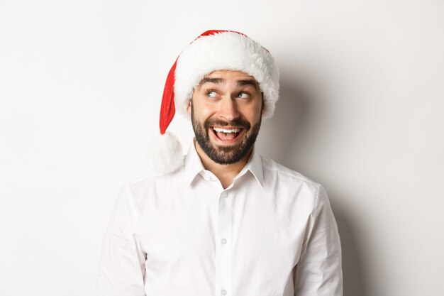 Close-up of happy bearded man in santa hat, celebrating christmas and New Year and looking at upper left corner, imaging something. white background.