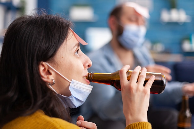 Close up of happy cheerful woman drinking beer while playing name game with multi ethnic friends having sticky notes on forehead wearing face mask keeping social distancing. Conceptual image.