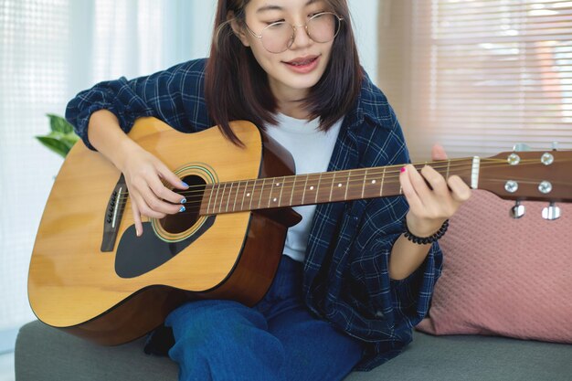 Крупным планом счастливые очки Азиатская девушка играет на акустической гитаре в гостиной дома Концепция отдыха дома