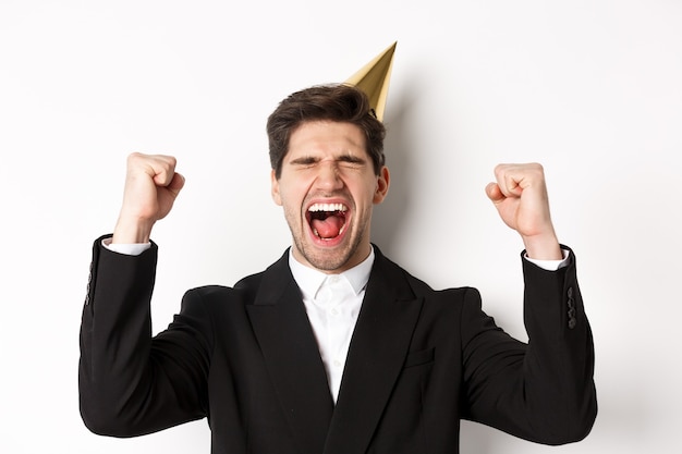 Close-up of happy good-looking man, wearing party hat and suit, raising hands up and rejoicing, celebrating new year, standing against white background.