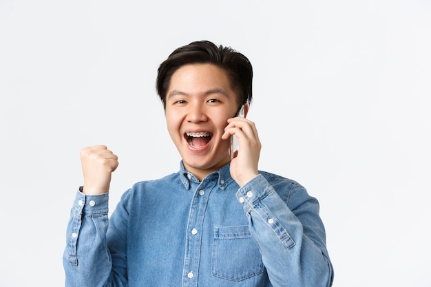 멋진 소식을 기뻐하는 행복한 미남 아시아 남자의 클로즈업, 휴대 전화와 주먹 펌프, 승리 및 예, 목표 달성. 남자는 고용주로부터 전화를 받고 일자리를 얻었으며 흰색 배경